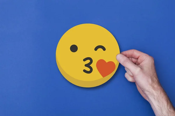 Mão masculina segurando um emoji emoticon beijando ícone de cabeça sorridente — Fotografia de Stock