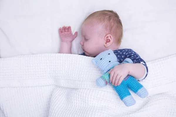 A cute young baby boy asleep under a blanket hugging a knitted teddy bear — Stock Photo, Image