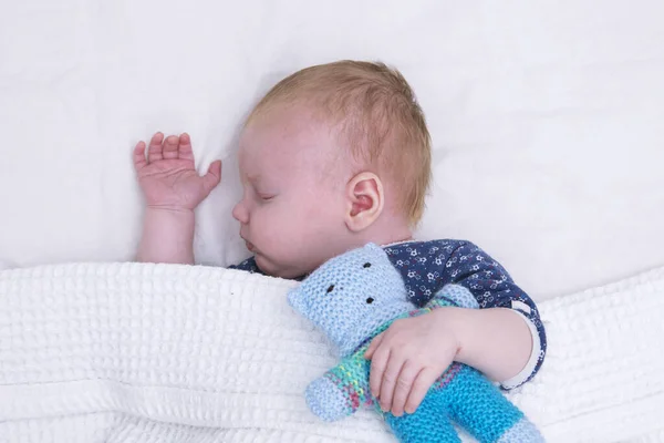 A cute young baby boy asleep under a blanket hugging a knitted teddy bear — Stock Photo, Image