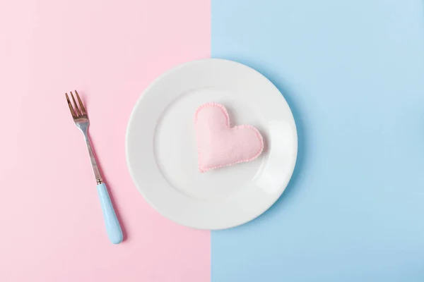 Felt heart on plate with fork on pastel pink and blue background for Valentines day romantic congratulation banner or cooking with love theme concept - flat lay of hand made decoration.