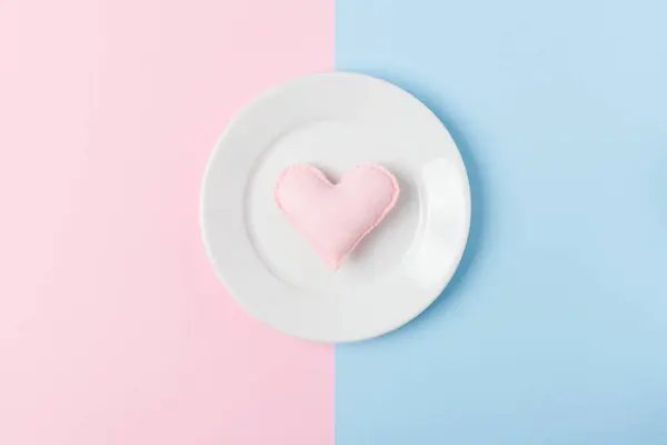 Felt heart on white plate on pastel pink and blue background for Valentines day romantic congratulation banner or cooking with love theme concept - hand made decoration for cafe or restaurant.