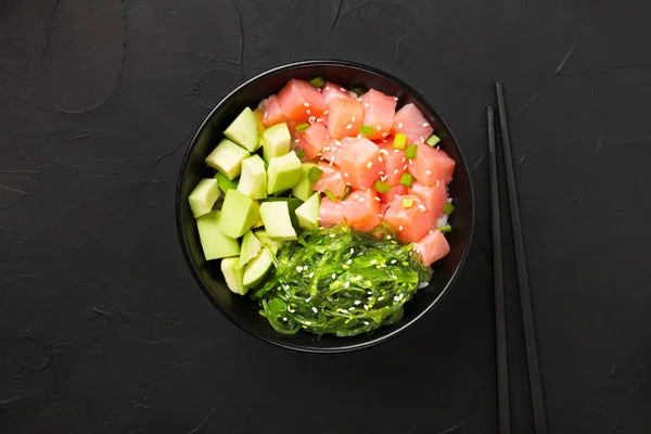 Poke with red fish, avocado and green seaweeds decorated with green onions and sesame seeds in bowl.