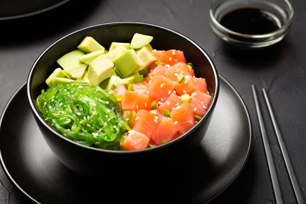 Poke with diced red fish, avocado and green seaweeds salad decorated with green onions and sesame seeds in black bowl with chopsticks and soy sauce on dark background - close up seafood photography.