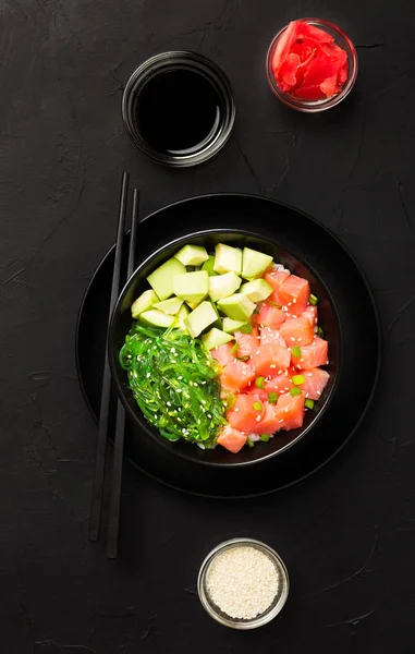 Top view of poke with diced red fish, avocado and green seaweeds salad decorated with green onions and sesame seeds in black bowl with soy sauce and ginger on dark background.