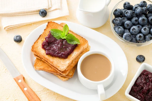 Fresh toasted bread with blueberry jam, raw ripe wild berries and cup of coffee with milk on yellow textured background.