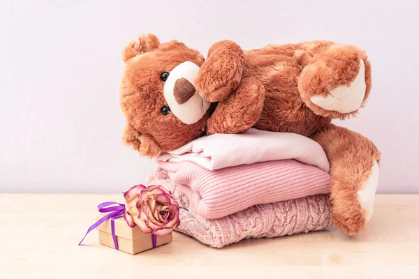 Teddy bear lies on Stack of winter or autumn womens clothes. Pile of knitted cozy warm pink sweaters or pullover and gift box with rose on wooden table.