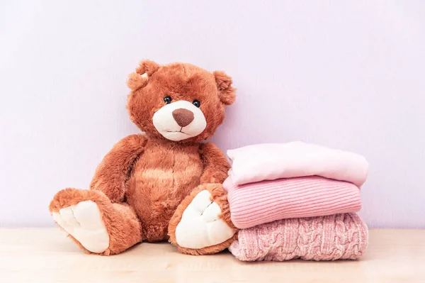 Teddy bear sits near the stack of winter or autumn womens clothes. Pile of rose knitted cozy warm pink sweaters or pullover on wooden table.