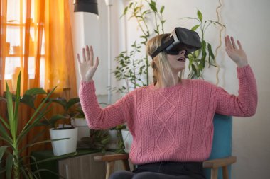 Kadın pembe kazaklı evde modern kullanarak sanal gerçeklik gözlükleri tadını koltuk etrafında arıyorsunuz ve hareketleri el ile kullanarak