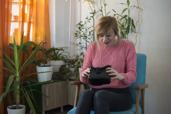 woman in pink sweater at home enjoying using virtual reality goggles in modern armchair Looking around and using gestures with hands