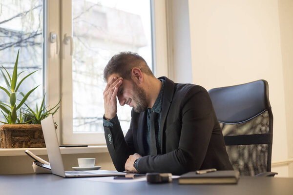 бизнесмен работает на ноутбуке в офисе головная боль у окна черный стол с ноутбуком бумаги телефон мигрень
