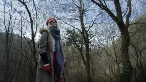 年轻迷人的女人与红帽徘徊在森林年轻迷人的女人与红色的帽子和红色的裙子徘徊沿着路径领先的槽茂密的森林 女性在森林中行走在冬天 — 图库视频影像
