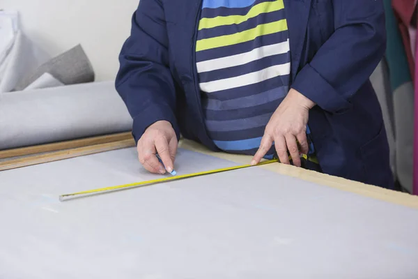 Closeup of an older woman in a furniture factory who is measuring and marking a grey material for the sofa with a chock.