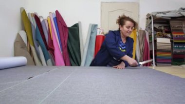 Karanlık paltolu yaşlı bir kadın kanepe için işaretli gri malzeme keser. Belgili tanımlık are arkasında malzemelerin farklı türleri. O ahşap bir masa üzerine malzeme kesim ve dikiş için hazırlanıyor.