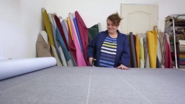 Karanlık paltolu yaşlı bir kadın kanepe için işaretli gri malzeme keser. Belgili tanımlık are arkasında malzemelerin farklı türleri. O ahşap bir masa üzerine malzeme kesim ve dikiş için hazırlanıyor.