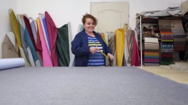 Karanlık bir kat büyük bir kadında ölçme ve kesme için gri malzeme hazırlar. Arkasında malzemeler farklı türleri vardır. O malzeme ahşap bir masa üzerine yayılır ve işleme için hazırlar. 