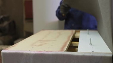 Mobilya üretim süreci. Genç ceket bir adam, bir karanlık bir köpük koltukta çalışma masası ahşap parçası üzerinde geçerlidir. Arkasında bir duvar bir yeri vardır araç ve Gereçleri. 