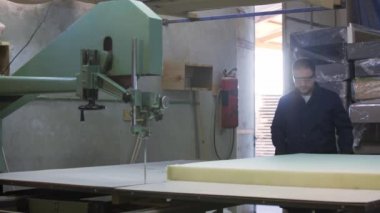 Mobilya üretim süreci. Genç ceket bir adam, bir karanlık kanepe için köpük kesme makinesi üzerinde kesti. Onun arkasında bitmiş kanepeler bir yeri vardır. Koruyucu gözlük takıyor ve eldiven.