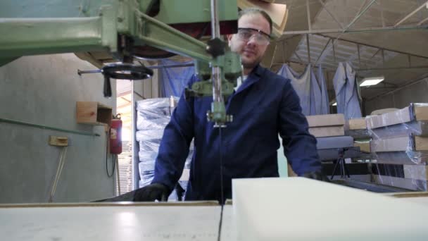 Processus de production de meubles. Un jeune homme vêtu d'un manteau sombre a coupé la mousse pour le canapé sur une machine à couper. Derrière lui, il y a beaucoup de canapés finis. Il porte des lunettes de sécurité et des gants. .
