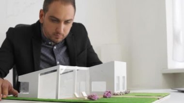 Ofiste iş adamı masada oturan bir ev modeline bakıyor. Düşünceli mimar proje hakkında düşünme. Etrafında hareketli parçalar, çatı ve yüzme havuzu ile oynuyor.