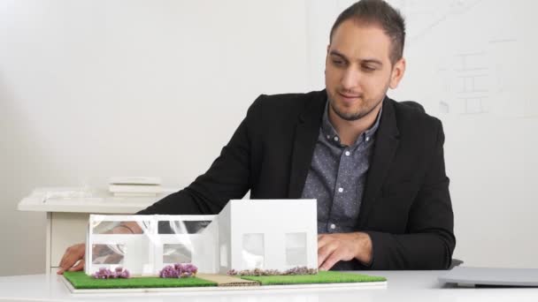 办公室里的商人坐在办公桌前看着一个房子的模型 有思想的建筑师对项目的思考 — 图库视频影像