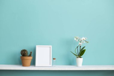 Resim çerçevesi mockup ile modern oda dekorasyonu. Saksı orkide ve kaktüs bitki ile pastel turkuaz duvara karşı beyaz raf.