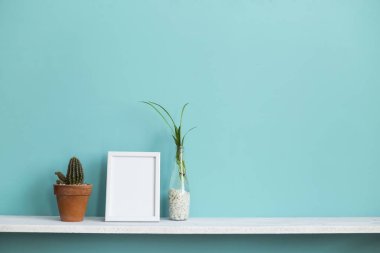 Resim çerçevesi mockup ile modern oda dekorasyonu. Su ve kaktüs örümcek bitki kesimleri ile pastel turkuaz duvara karşı beyaz raf.
