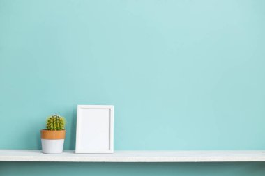 Resim çerçevesi mockup ile modern oda dekorasyonu. Saksılı kaktüs bitkisi ile pastel turkuaz duvara karşı beyaz raf.