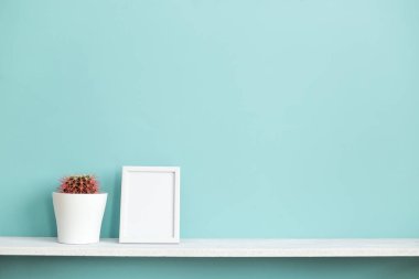 Resim çerçevesi mockup ile modern oda dekorasyonu. Saksılı kaktüs bitkisi ile pastel turkuaz duvara karşı beyaz raf.