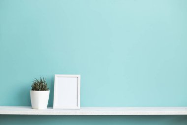 Resim çerçevesi mockup ile modern oda dekorasyonu. Saksı lı sulu bitki ile pastel turkuaz duvara karşı beyaz raf.
