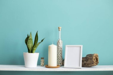 Resim çerçevesi mockup ile modern oda dekorasyonu. Mum ve şişe kayalar ile pastel turkuaz duvara karşı beyaz raf. Saksıyılan bitkisi