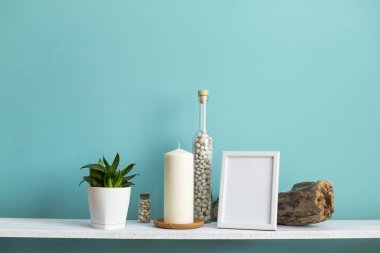 Resim çerçevesi mockup ile modern oda dekorasyonu. Mum ve şişe kayalar ile pastel turkuaz duvara karşı beyaz raf. Saksıyılan bitkisi