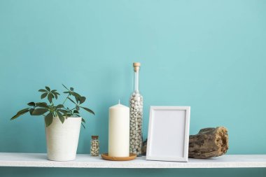 Resim çerçevesi mockup ile modern oda dekorasyonu. Mum ve şişe kayalar ile pastel turkuaz duvara karşı beyaz raf. Saksılı schefflera bitkisi