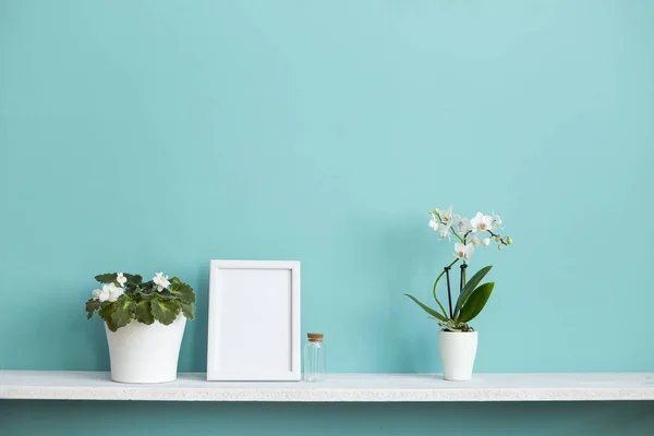 Moderna decoración de la habitación con marco de imagen maqueta. Estante blanco contra pared de color turquesa pastel con orquídea en maceta y planta violeta . — Foto de Stock