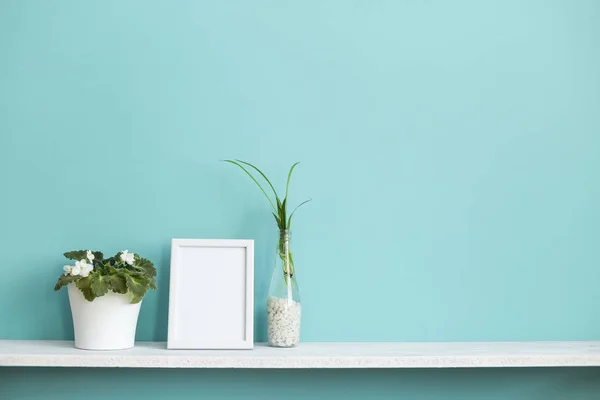 Resim çerçevesi mockup ile modern oda dekorasyonu. Su ve menekşe örümcek bitki kesimleri ile pastel turkuaz duvara karşı beyaz raf.