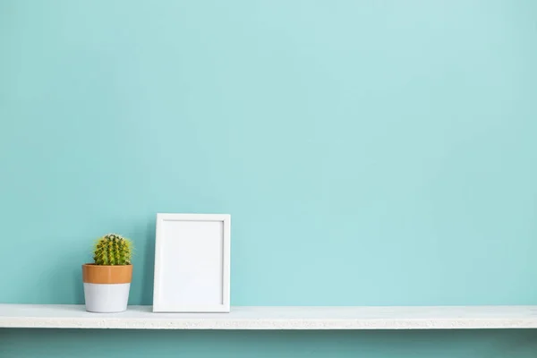 Resim çerçevesi mockup ile modern oda dekorasyonu. Saksılı kaktüs bitkisi ile pastel turkuaz duvara karşı beyaz raf.