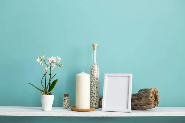 Moderna decoración de la habitación con marco de imagen maqueta. Estante blanco contra pared de color turquesa pastel con vela y rocas en botella. Planta orquídea en maceta — Foto de Stock