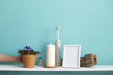 Resim çerçevesi mockup ile modern oda dekorasyonu. Mum ve şişe kayalar ile pastel turkuaz duvara karşı beyaz raf. El saksı menekşe bitki aşağı koyarak