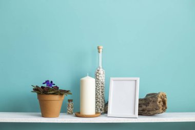 Resim çerçevesi mockup ile modern oda dekorasyonu. Mum ve şişe kayalar ile pastel turkuaz duvara karşı beyaz raf. Saksılı menekşe bitkisi