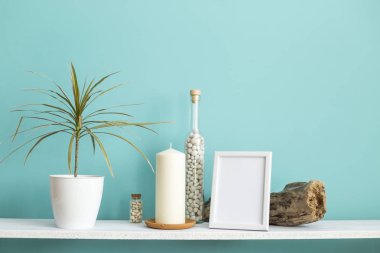 Resim çerçevesi mockup ile modern oda dekorasyonu. Mum ve şişe kayalar ile pastel turkuaz duvara karşı beyaz raf. Saksılı dracaens bitki