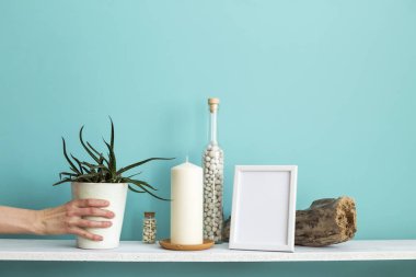 Resim çerçevesi mockup ile modern oda dekorasyonu. Mum ve şişe kayalar ile pastel turkuaz duvara karşı beyaz raf. El saksı sulu bitki aşağı koyarak