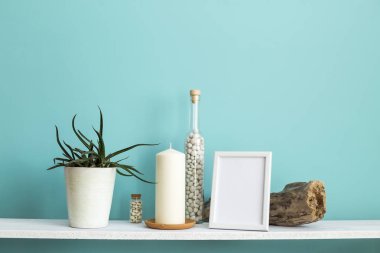 Resim çerçevesi mockup ile modern oda dekorasyonu. Mum ve şişe kayalar ile pastel turkuaz duvara karşı beyaz raf. Saksılı sulu bitki