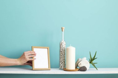 Resim çerçevesi mockup ile modern oda dekorasyonu. Mum ve şişe kayalar ile pastel turkuaz duvara karşı beyaz raf. Saksılı sulu bitki