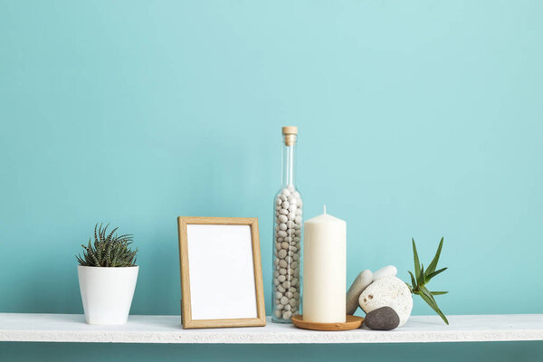 Modern room decoration with Picture frame mockup. White shelf against pastel turquoise wall with Candle and rocks in bottle. Potted succulent plant