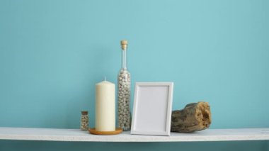 Resim çerçevesi mockup ile modern oda dekorasyonu. Mum ve şişe kayalar ile pastel turkuaz duvara karşı beyaz raf. Saksıyılan bitkisi aşağı koyarak El