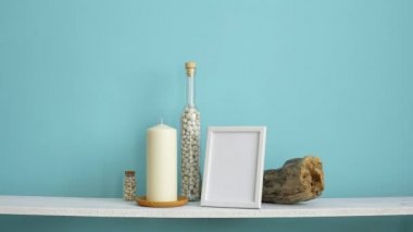 Resim çerçevesi mockup ile modern oda dekorasyonu. Mum ve şişe kayalar ile pastel turkuaz duvara karşı beyaz raf. Saksıyılan bitkisi aşağı koyarak El