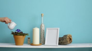 Resim çerçevesi mockup ile modern oda dekorasyonu. Mum ve şişe kayalar ile pastel turkuaz duvara karşı beyaz raf. El sulama saksı menekşe bitki