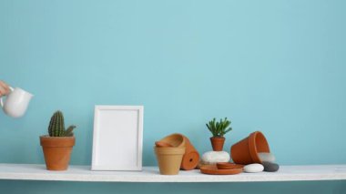 Resim çerçevesi mockup ile modern oda dekorasyonu. Çanak çömlek ve etli bitki ile pastel turkuaz duvara karşı beyaz raf. El sulama saksı kaktüs bitki.