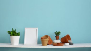 Resim çerçevesi mockup ile modern oda dekorasyonu. Çanak çömlek ve etli bitki ile pastel turkuaz duvara karşı beyaz raf. El sulama saksı yılan bitki.