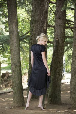 Çam ağacının altında dururken siyah elbise giyen güzel yalınayak genç kadınların tam uzunlukta portre