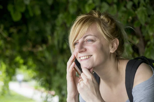 公園のベンチに座り、電話をかけて座っている女性の肖像画。彼女は微笑んでいる. — ストック写真